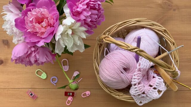 Video of needlework. Process of hand crochet a lace square doily made of cotton and acrylic yarn. Vase with fresh pink peonies and basket with threads and hooks on a wooden table