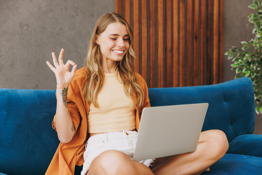 Young IT woman wears brown shirt hold use work on laptop pc computer sit on blue sofa couch stay at home hotel flat relax spend free spare time in living room indoors grey wall. People lounge concept.