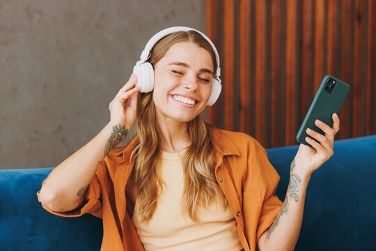 Young happy woman she wear brown casual clothes headphones listen to music hold use mobile cell phone close eyes sit on blue sofa indoor rest at home in own room apartment. People lifestyle concept..