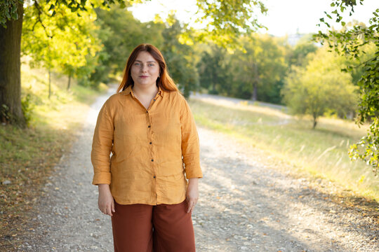 Full length front view portrait of a calm young Caucasian woman with red hair and glasses standing on a path in a green sunny forest or public park.