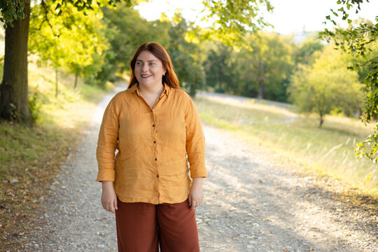 Full length front view portrait of a calm young Caucasian woman with red hair and glasses standing on a path in a green sunny forest or public park.