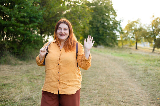 A front view portrait of a happy young female tourist with long red hair and an orange shirt waving her hand in greeting while hiking with a backpack in a green forest.