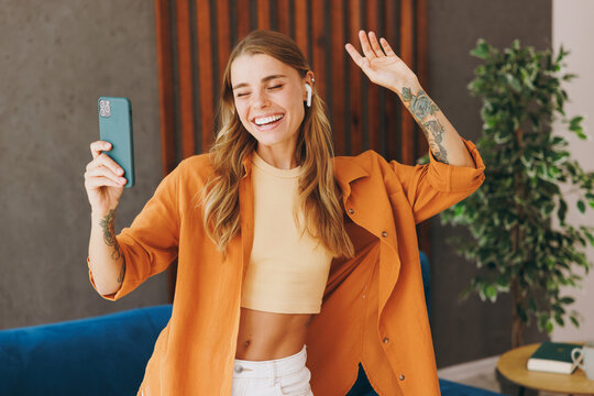 Young fun cool woman wear brown shirt listen music in earphones use mobile cell phone dancing near blue sofa stay at home flat relax spend free spare time in living room indoor. People lounge concept