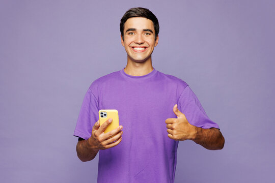 Young smiling Caucasian man he wear violet t-shirt casual clothes hold in hand use mobile cell phone show thumb up isolated on plain pastel light purple background studio portrait. Lifestyle concept.