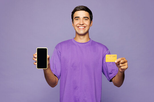 Young happy man he wears violet t-shirt casual clothes using blank screen mobile cell phone hold credit bank card doing online shopping order delivery booking tour isolated on plain purple background.