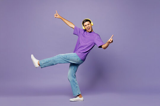 Full body young happy man he wear violet t-shirt casual clothes listen to music in headphones raise up hands dance isolated on plain pastel light purple background studio portrait. Lifestyle concept.