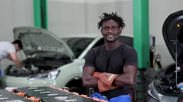 Auto technicians in a workshop servicing electric vehicles, with a large battery pack on a table and open hoods. Mechanic in blue overalls smiling, colleague working in background.