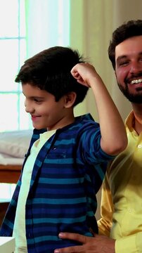 Indian Asian young female dentist demonstrating a denture model to a small boy sitting on his father&rsquo;s lap in modern clinic, as the cheerful child flexes his muscle to show strong teeth, all smiling