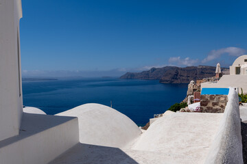 Obraz premium Traditional Cycladic architecture with smooth white domed roofs on a cliffside in Oia, Santorini. Deep blue sea and distant volcanic islands visible under a clear sky on a sunny day.