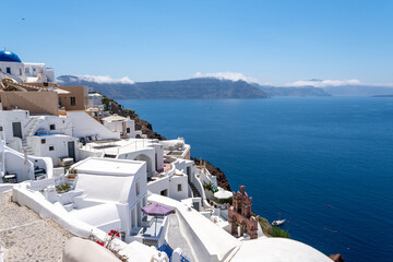 Obraz premium Traditional white Cycladic houses cascading down the cliffside in Oia, Santorini. Blue church dome, pink bell tower, and calm Aegean Sea with volcanic islands under clear sky.