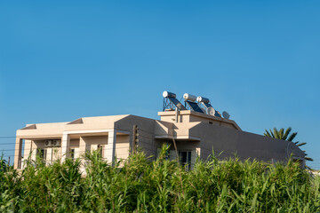 Two-story beige residential house with solar panels and water heating systems on the rooftop. Green reed plants in foreground and clear blue sky background on a sunny day. © Pasha