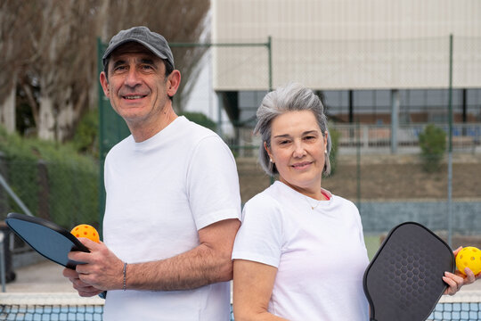 Active senior couple smiling on a pickleball court, holding paddles and balls, ready to play