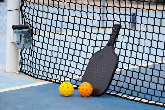 Pickleball paddle and two balls leaning against the net on a blue outdoor court