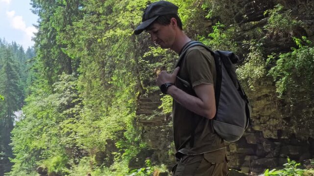 A horizontal tracking profile shot of a male hiker with a backpack walking past a steep mossy rock wall in a lush summer forest, ideal for adventure marketing and outdoor lifestyle branding.