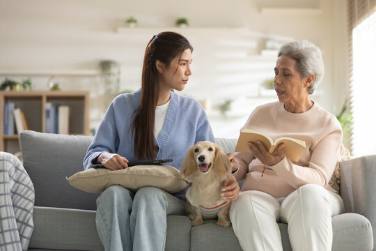 Serious young woman and senior mother talking on living room sofa with pet dog. Caring daughter listening to elderly parent reading book. They look concerned and focused on conversation together