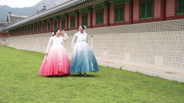 Two women in flowing traditional dresses stroll together. They move along a manicured green lawn. Ancient stone walls and red-tiled roofs frame them. Their expressions convey calm, gentle joy