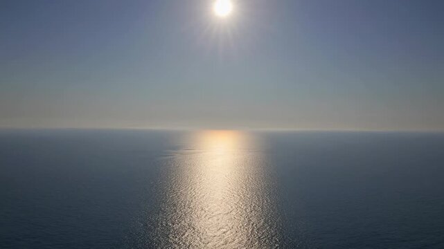 Bright sun shines over calm ocean waters, reflecting light across the surface, creating a serene atmosphere in a clear blue sky above the horizon
