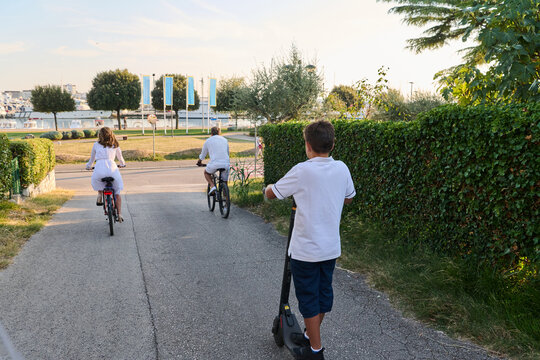 Family Riding Bicycles and Electric Scooter Along Seaside Path Near Marina in Summer Morning