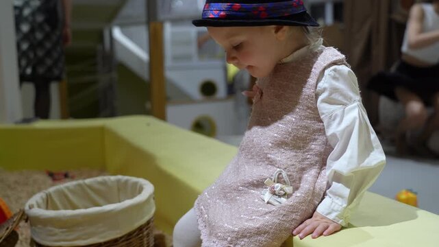 Stylish toddler in tweed outfit and patterned hat sits near sensory bin, looking towards unseen elements.