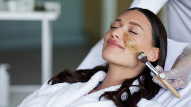 Happy woman enjoying soothing facial skincare treatment as beauty specialist applying nourishing cosmetic mask in modern wellness salon, panorama, free space