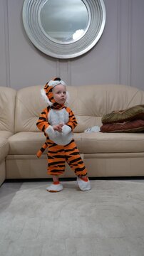 Cute toddler girl in orange tiger costume standing and playing in living room, wearing plush striped outfit with hood and tail, having fun during dress up time at home