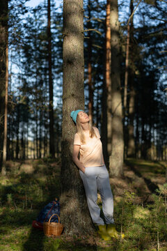 Tranquil smiling woman forager backpacker taking moment to enjoy harmony of nature in forest, smiling relaxed female leaning against tree in summer woodlands during mushroom picking hobby activity.