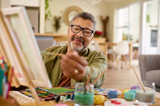 Satisfied multiethnic senior man painting on canvas at home