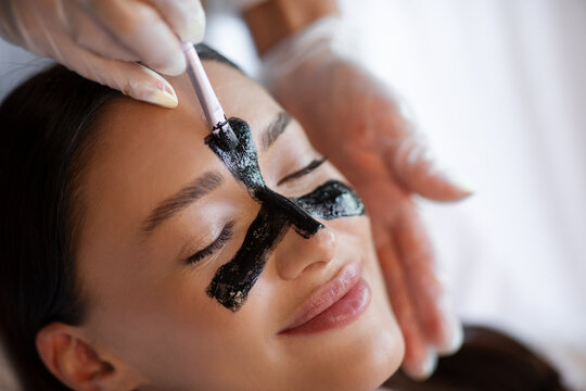 Closeup of relaxed woman receiving charcoal face mask on T-zone while esthetician performing luxury skincare procedure in beauty center