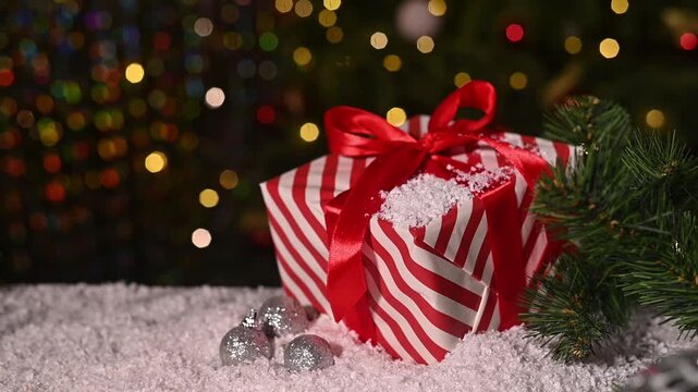 Christmas gift wrapped with red ribbon, soft bokeh lights from tree create cozy holiday atmosphere. Winter present with red ribbon, Christmas tree lights blur softly in background. New Year backdrop.