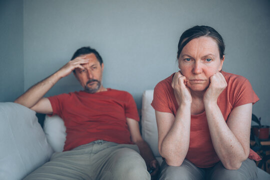 Upset middle-aged couple sits apart on a couch after an argument, showing emotional distance, frustration and stress in a strained marriage at home.