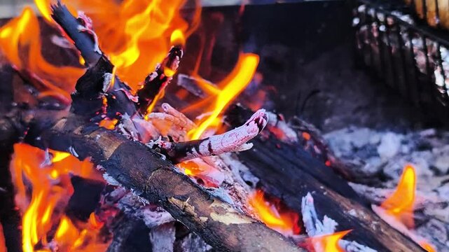 Fire in grill with burning wood and hot coals closeup, barbecue concept. Fire flames flicker and spread across logs, fire produces heat and light, outdoor cooking atmosphere and energy of combustion