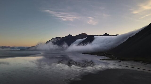 Aerial tracking across black sand tidal flats mirroring the Vestrahorn ridge and its fog waterfall, Iceland