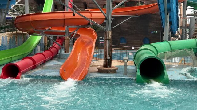  Colorful blue, orange, green, and red water slides twist into a pool inside an indoor water park with a wooden dome roof and palm trees.