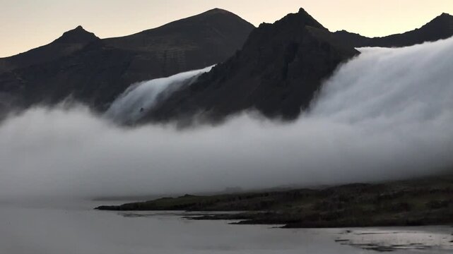 Drone skims above black tidal flats toward Vestrahorn as fog cascades down the ridge at dawn, Stokksnes, Iceland