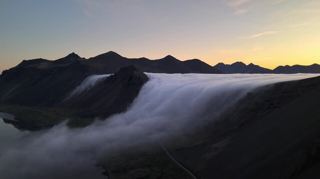 High aerial sweep along the Ring Road as fog inversion pours over the mountain ridge at sunrise, East Iceland