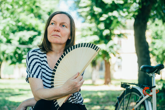 Middle-aged woman cooling herself with a handheld fan while sitting on a park bench during a hot summer day. Heatwave, outdoor comfort and urban lifestyle concept.