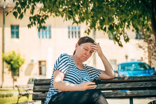 Middle-aged woman sitting on park bench using smartphone and relaxing in urban summer setting,