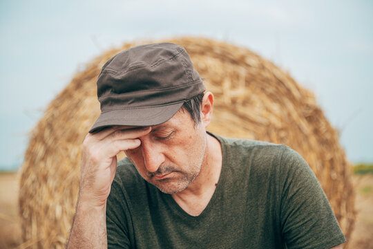 Worried agricultural worker in a cap against a straw bale on a field, reflecting on crop failure or financial loss.