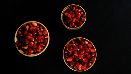 Fototapeta premium Wild strawberries are arranged in three ceramic bowls. Showcasing their vibrant red color and fresh appearance against a deep black background from a top down perspective