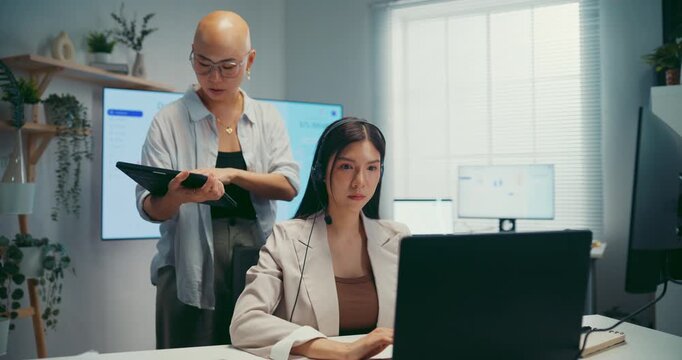 Young Asian businesswoman hold tablet to mentor trainee in call center workspace office. Corporate customer service training, employee support coach, telemarketing agent, teamwork help desk concept.