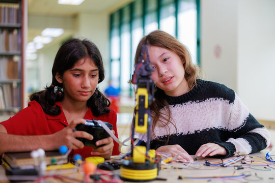 Two focused teenage female students collaborate on building and programming a yellow robotic arm with electronic components during a STEM education project in a school library.