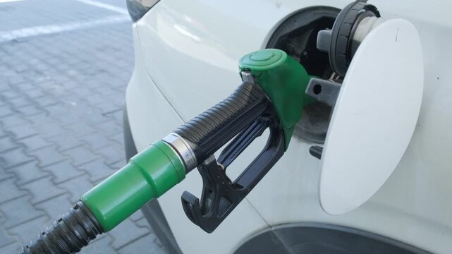 A horizontal close up shot of a green petrol pump nozzle fueling a white crossover vehicle at a gas station, perfect for automotive services, travel logistics, and energy industry marketing.