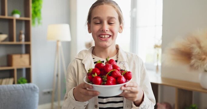 Smiling child holding bowl of strawberries at home. Fresh fruit snack suggests natural nutrition and wholesome eating in a cozy living room with daylight. Ideas for healthy home snacking.