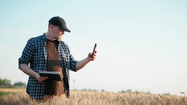 Farmer inspecting a ripe wheat field while using a smartphone and taking notes in a notebook, capturing modern agriculture, crop monitoring and farm management.