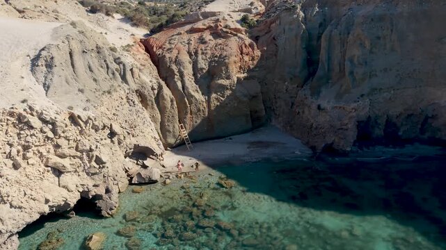 Experience breathtaking views of Milos in Greece, where crystal clear waters meet majestic cliffs. A couple enjoy a serene day at the A couple of men and woman walks at the Tsigrado Beach