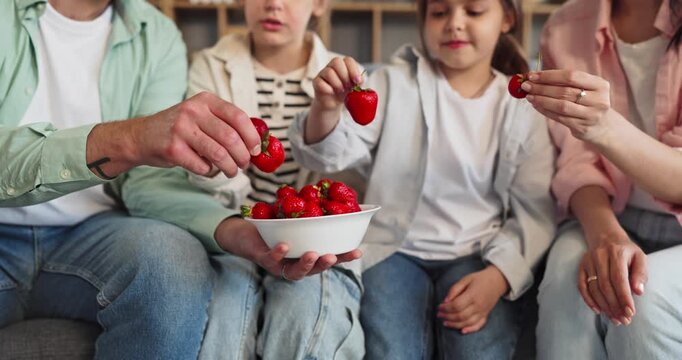 Family children and parents eating strawberries together. Close view of hands taking a bowl showing fresh fruit and healthy snack sharing at home. Concept shows family happiness while sharing.