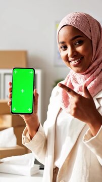 Smiling young woman in hijab showing smartphone with green screen, pointing to it, in a bright office setting, vertical shot.