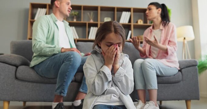 Child parents argument conflict at home, crying. A young girl covers her face on the floor as parents argue on the sofa, showing family stress and trauma. Family conflict and trauma theme.