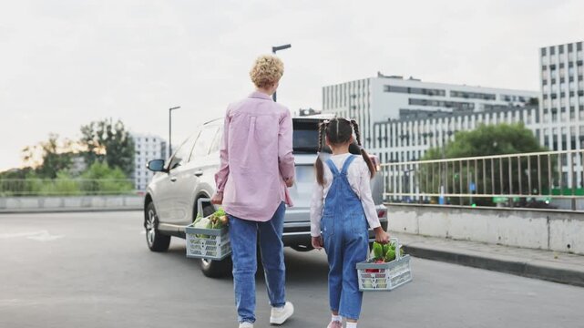 A Caucasian mother and her young daughter are packing groceries into the trunk of a car after a shopping trip