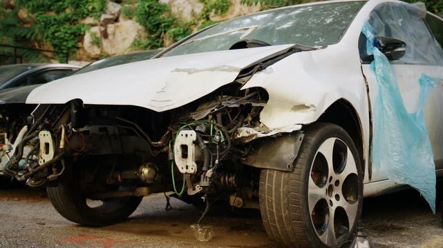 Severely damaged white car with missing front bumper and exposed mechanical parts parked in a scrapyard